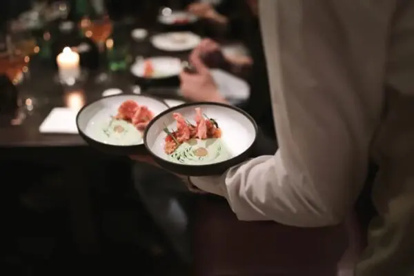 A server carrying two plated crudo dishes through the candlelit dining room at Brugada Kitchen & Bar
